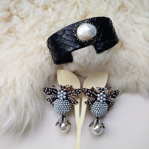 Może przedstawiać: Czarna skórzana bransoletka z dużą białą perłą i czarnymi kryształami. Dwie pasujące kolczyki w kształcie pszczoły z białymi perłami i dużą białą perłą zwisającą poniżej.