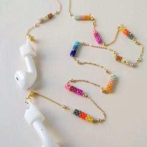 Könnte beinhalten: Eine goldene Kette mit bunten Perlenabschnitten verbindet zwei weiße drahtlose Ohrhörer. Die Perlen sind in einem Farbverlauf von rosa, orange, gelb, grün, blau und lila angeordnet.
