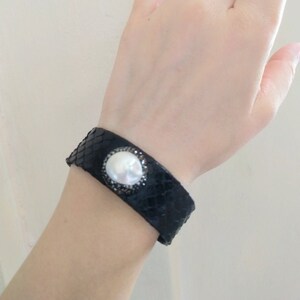 May include: A black leather bracelet with a large white pearl and black rhinestones.