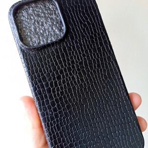Può includere: Una custodia per smartphone nera caratterizzata da un motivo testurizzato a pelle di coccodrillo sulla superficie posteriore. La custodia include un ritaglio quadrato rialzato per la fotocamera nell'angolo in alto a sinistra con un interno scuro.