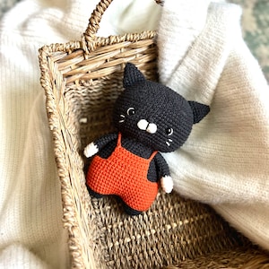 May include: A handmade black crochet cat wearing orange overalls, white paws, and a white muzzle. The cat is in a woven basket with a handle, resting on a white sweater. The cat has small white whiskers and a smiling expression.