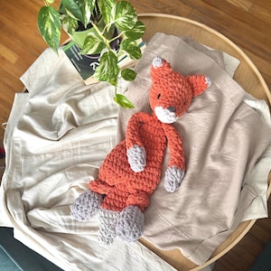 May include: A crocheted fox plush toy with orange fur and gray accents. The toy is lying on a white blanket.
