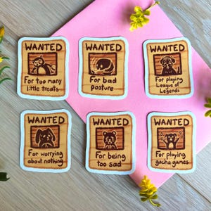 Pode incluir: Seis autocolantes retangulares com o tema "WANTED". Cada um apresenta uma ilustração de animal de desenho animado e uma razão humorística para ser "procurado", como "Por demasiadas guloseimas" ou "Por jogar League of Legends". Os autocolantes estão numa superfície rosa.