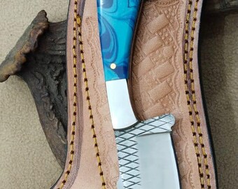 May include: A handcrafted knife with a blue and white marbled handle and a serrated blade, nestled in a custom-made leather sheath. The sheath is tan with decorative stitching and embossed details.
