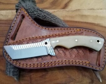 May include: A handmade knife with a cream-colored handle and a silver blade rests in a brown leather sheath. The sheath is intricately stitched and embossed with a decorative pattern. The knife blade has a unique, angled shape.