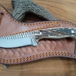 May include: A custom hunting knife with a serrated blade and a handle made of antler material. The knife rests in a brown leather sheath with decorative stitching and embossed patterns. The overall design suggests a handcrafted, outdoor-themed product.