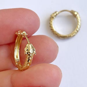 May include: Gold-colored hoop earrings with a detailed snake design. One earring is in focus, held by a hand, while another earring is in the background. The earrings have a textured surface and a secure clasp.