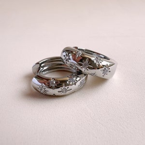 Può includere: Due orecchini a cerchio color argento con un design a stella. Ogni orecchino presenta molteplici abbellimenti a forma di stella con una piccola pietra trasparente al centro. Gli orecchini hanno una finitura liscia e lucida e una forma arrotondata.