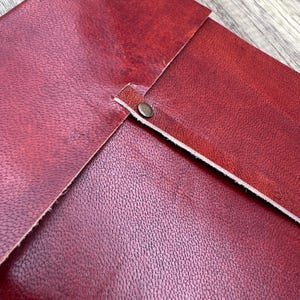 May include: A close-up of a burgundy leather journal with a flap closure and a metal clasp. The journal has a textured surface and a white stitching detail.