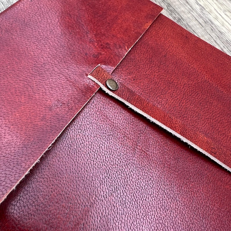 May include: A close-up of a burgundy leather journal with a flap closure and a metal clasp. The journal has a textured surface and a white stitching detail.