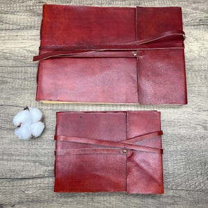 May include: Two burgundy leather journals with straps and metal clasps. The larger journal is on top of the smaller journal.