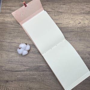May include: An open, blank, cream-colored journal with a pink leather cover and a brown leather strap. The journal is lying open on a wooden surface.