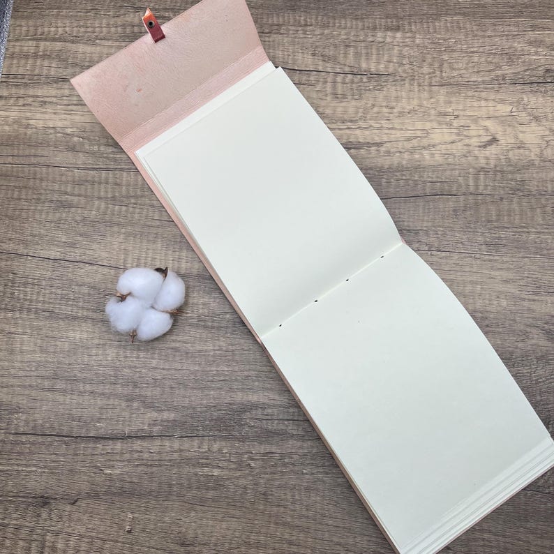 May include: An open, blank, cream-colored journal with a pink leather cover and a brown leather strap. The journal is lying open on a wooden surface.