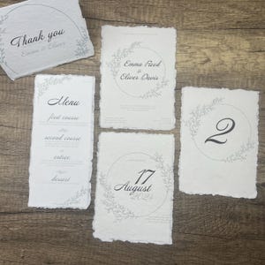 May include: Wedding stationery set with a rustic theme. The set includes a thank you card, a menu card, a save the date card, and a table number card. Each card features a delicate floral wreath design in gray and white.