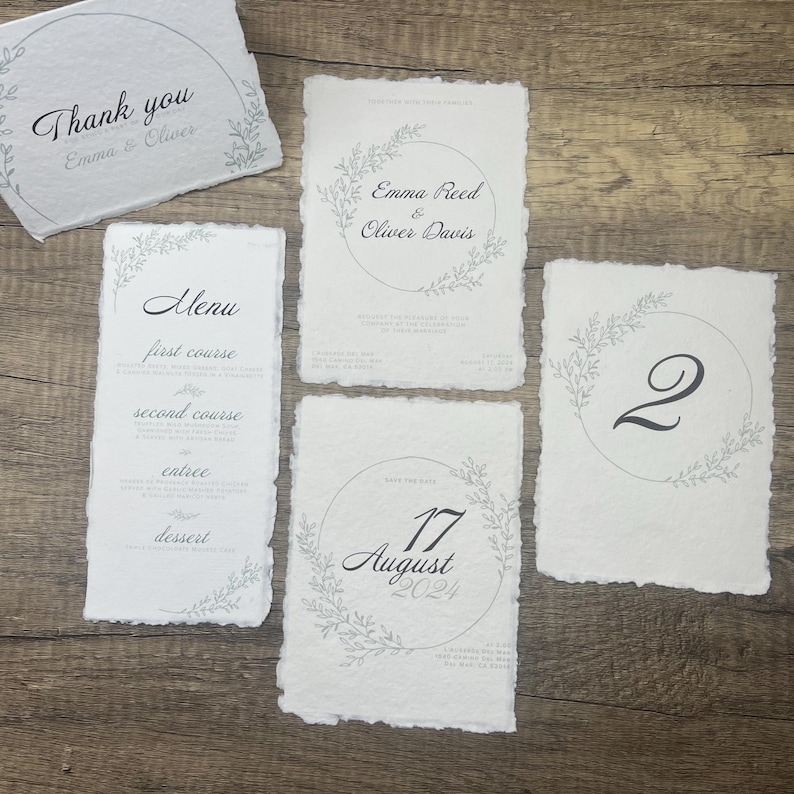 May include: Wedding stationery set with a rustic theme. The set includes a thank you card, a menu card, a save the date card, and a table number card. Each card features a delicate floral wreath design in gray and white.