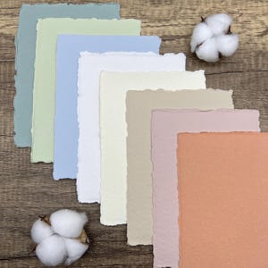 May include: A set of ten textured paper sheets in various shades of blue, green, white, beige, pink, and orange. The paper sheets are arranged in a fan shape on a wooden surface.