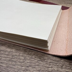 May include: A close-up of a leather-bound journal with a stack of blank pages. The journal is open and the pages are visible. The leather cover is a light pink color.