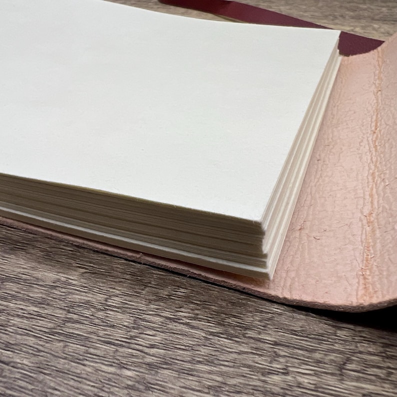 May include: A close-up of a leather-bound journal with a stack of blank pages. The journal is open and the pages are visible. The leather cover is a light pink color.