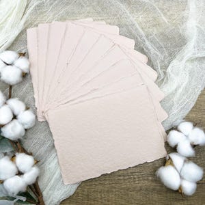 May include: A stack of pale pink handmade paper sheets with deckled edges. The paper has a textured surface and is arranged in a fan shape. The paper is on a white fabric background.