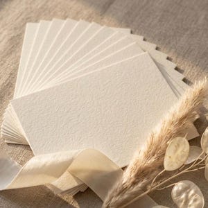 May include: A stack of rectangular, off-white cards with a textured, heavy-weight paper finish. The cards are arranged in a fan shape. A light-colored silk ribbon and a dried, feathery botanical stem are positioned across the bottom of the cards.