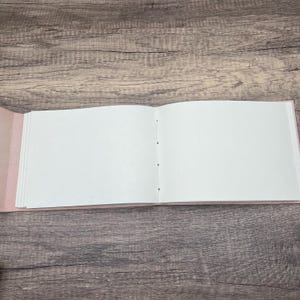 May include: A blank, white, open book with a pink cover. The book is lying flat on a wooden surface.