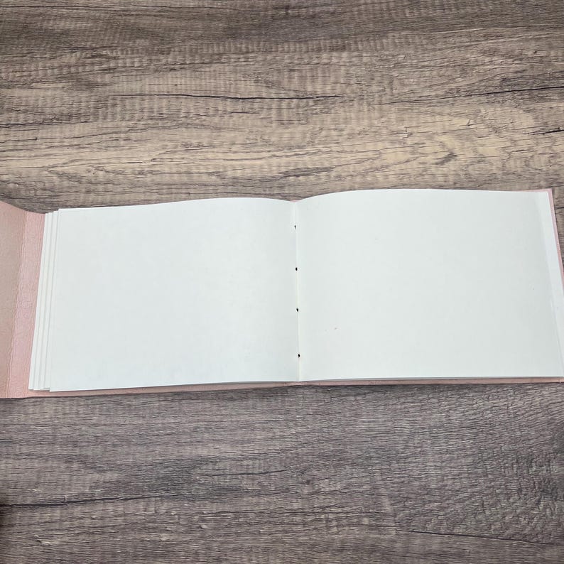 May include: A blank, white, open book with a pink cover. The book is lying flat on a wooden surface.
