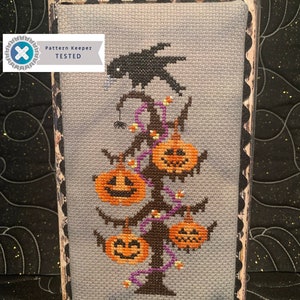 May include: A cross-stitch pattern of a spooky Halloween tree with a black crow perched on top. The tree is decorated with orange pumpkins and purple lights. The pattern is framed in a black wood frame with a scalloped edge. The text "Pattern Keeper TESTED" is visible in the lower left corner.
