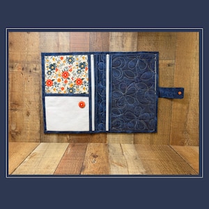 May include: A fabric organizer wallet featuring a dark blue quilted exterior with a floral pattern on the left interior panel, a white pocket with an orange button, two vertical white zippers, and a side tab with an orange snap closure.