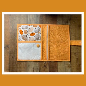May include: Open, quilted fabric organizer in shades of orange and white. The interior features a pumpkin print pocket, a white pocket, and a zippered compartment. The exterior is a solid orange with a quilted pumpkin design. A small orange button secures the organizer.