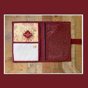 May include: Open burgundy book cover with a quilted design. The left side features a cream-colored map print with the text "The Marauders Map". A white pocket and a button closure are also visible. The cover is on a wooden surface.