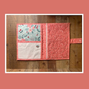 May include: Open, quilted fabric wallet in coral pink with a floral pattern. The interior features a light blue floral print, a white pocket, and a button closure. A white zipper runs down the center. The wallet is open and lying flat on a wooden surface.