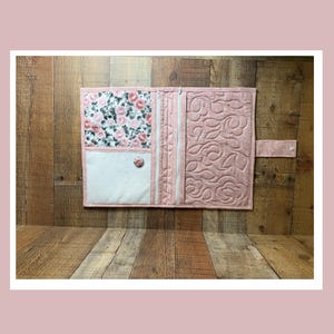 May include: A pink quilted journal cover with a floral print pocket and a button closure. The cover has a white pocket and a floral print on the left side, with a zipper and quilted design on the right side.