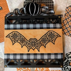 May include: A Halloween decoration with a black bat cross-stitch on an orange backdrop. It's framed in black wood, with black and white checked ribbon and a black velvet pumpkin for a festive touch.