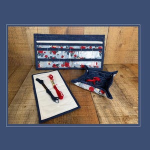 May include: A rectangular denim organizer with clear vinyl pockets featuring a red floral pattern, a matching square fabric tray with folded corners, and a rectangular white fabric mat holding two skeins of embroidery floss in black and red.