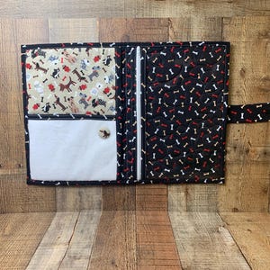May include: Open fabric wallet with a black background and a pattern of white and red dog bones. The interior features a beige fabric with various dog illustrations and a white pocket. A button closure secures the wallet.
