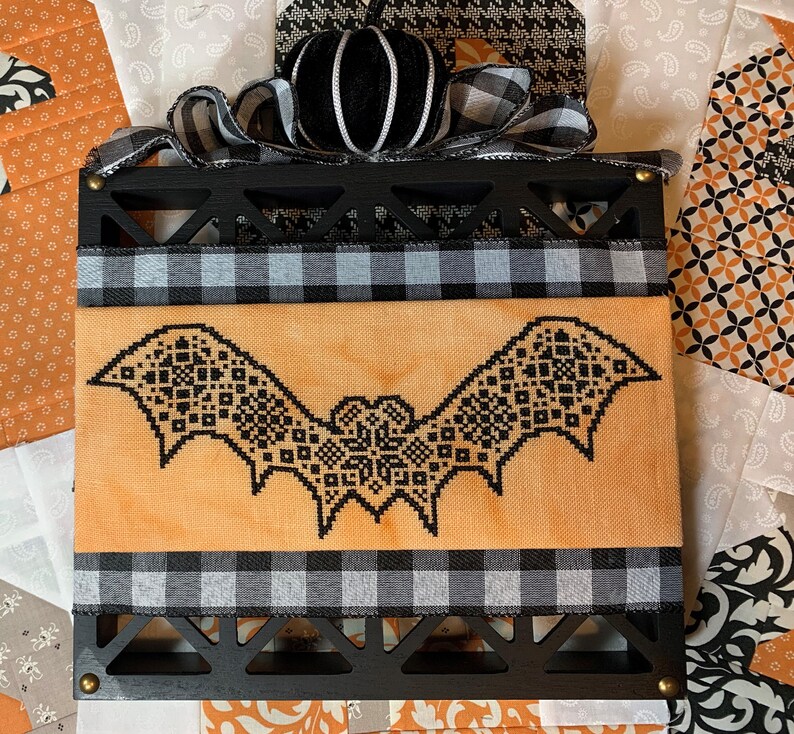 May include: A Halloween decoration with a black bat cross-stitch on an orange backdrop. It's framed in black wood, with black and white checked ribbon and a black velvet pumpkin for a festive touch.