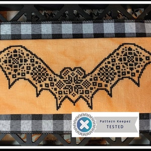May include: A cross-stitch bat design in black on an orange fabric, framed with a black wooden border and a black and white checkered ribbon. The image includes a "Pattern Keeper TESTED" logo, suggesting a craft project.