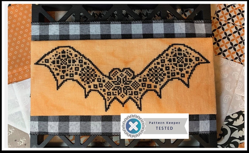 May include: A cross-stitch bat design in black on an orange fabric, framed with a black wooden border and a black and white checkered ribbon. The image includes a "Pattern Keeper TESTED" logo, suggesting a craft project.