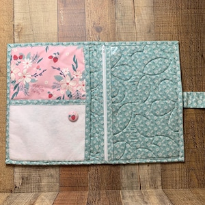 May include: Open quilted fabric journal cover with a floral design. The cover is teal with a white floral pattern and a pink floral pocket. It has a white zipper closure and a button closure.