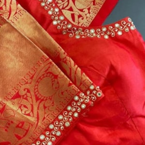 以下が含まれることがあります： 金色の刺繍と鏡の装飾が施された赤いシルクのブラウス。ブラウスはVネックで、金色の縁取りが施されています。