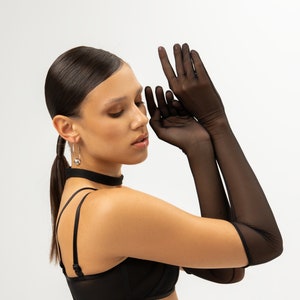 May include: A woman wearing a black bra and long black sheer gloves. She is looking down and touching her face with her hands.