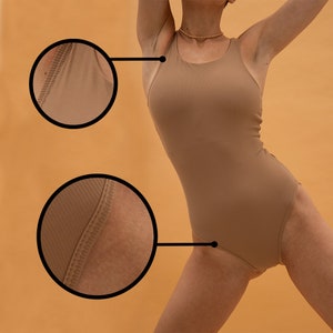 May include: A tan-colored bodysuit with a scoop neck and a racerback design. The fabric has a ribbed texture, and the seams are visible. The bodysuit is form-fitting and appears to be made of a stretchy material. The background is a solid orange color.