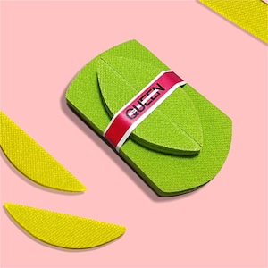 Può includere: Un oggetto a forma di foglia verde lime con una fascia rossa con la scritta "QUEEN" in bianco. L'oggetto è su uno sfondo rosa, con oggetti gialli a forma di foglia negli angoli.