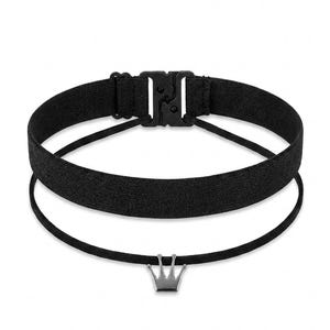 Może przedstawiać: Czarny naszyjnik typu choker ze srebrną zawieszką w kształcie korony. Choker posiada szeroką, teksturowaną opaskę z czarnym plastikowym zapięciem. Poniżej wisi cieńszy, sznurkowy naszyjnik z małym urokiem korony.