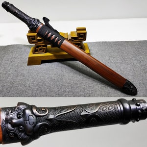 Könnte beinhalten: Ein schwarzes und braunes Schwert mit aufwendigen Schnitzereien am Griff und der Klinge. Das Schwert liegt auf einem Holzständer mit goldener Oberfläche.