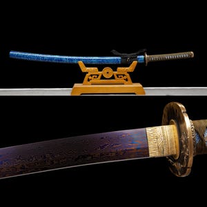 May include: A katana sword with a blue speckled scabbard resting on a wooden stand. The blade features a dark, wavy Damascus steel pattern with a gold-colored collar and a textured, dark-wrapped handle with a circular metal hand guard.