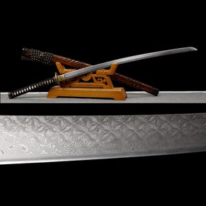 Pode incluir: Uma espada katana japonesa com uma lâmina curva, repousando sobre um suporte de madeira. A espada tem uma empunhadura preta e uma bainha decorativa com um padrão vermelho e marrom. A lâmina é prateada e tem um padrão detalhado.