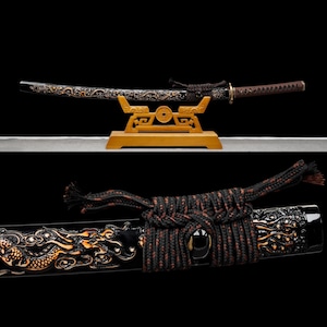 May include: A Japanese katana sword with a black scabbard and intricate gold dragon detailing. The sword rests on a wooden stand, showcasing the blade and handle. The handle is wrapped in a brown material, and the scabbard features a black and red cord.