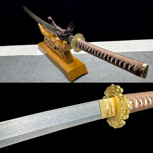 Pode incluir: Uma katana japonesa tradicional com um cabo trançado marrom e preto e uma tsuba de metal dourado. A espada é exibida em um suporte de madeira.