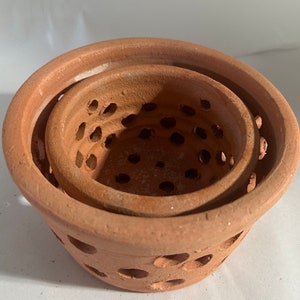 May include: Three terracotta pots of varying sizes nested together. Each pot has a series of evenly spaced holes around the sides. The pots are a warm, earthy brown color, and the texture appears slightly rough.